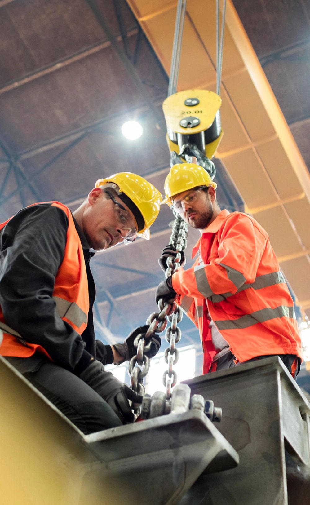 Two workers in hard hats and safety glasses rigging a chain hoist
